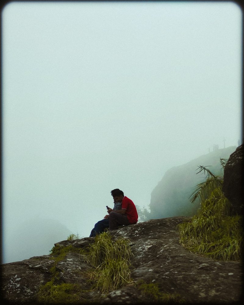 A person in a red top sitting on a rocky grassy hillside beneath mist