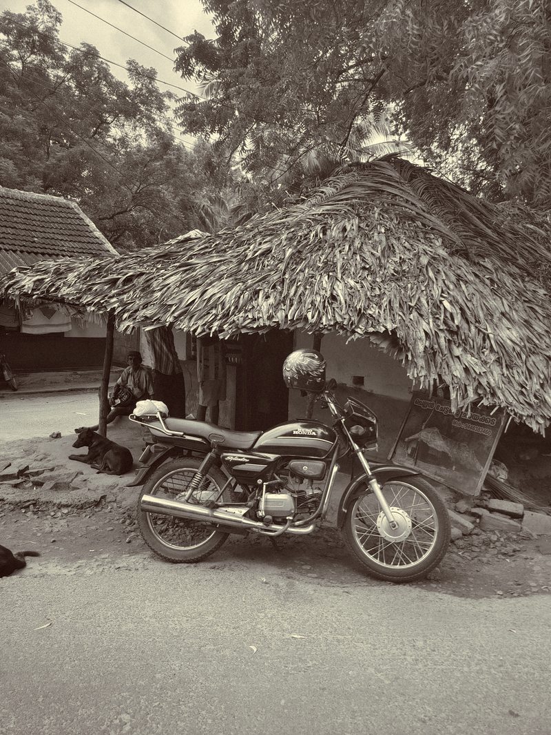 A motorcycle parked outside a thatched roadside building in monochrome