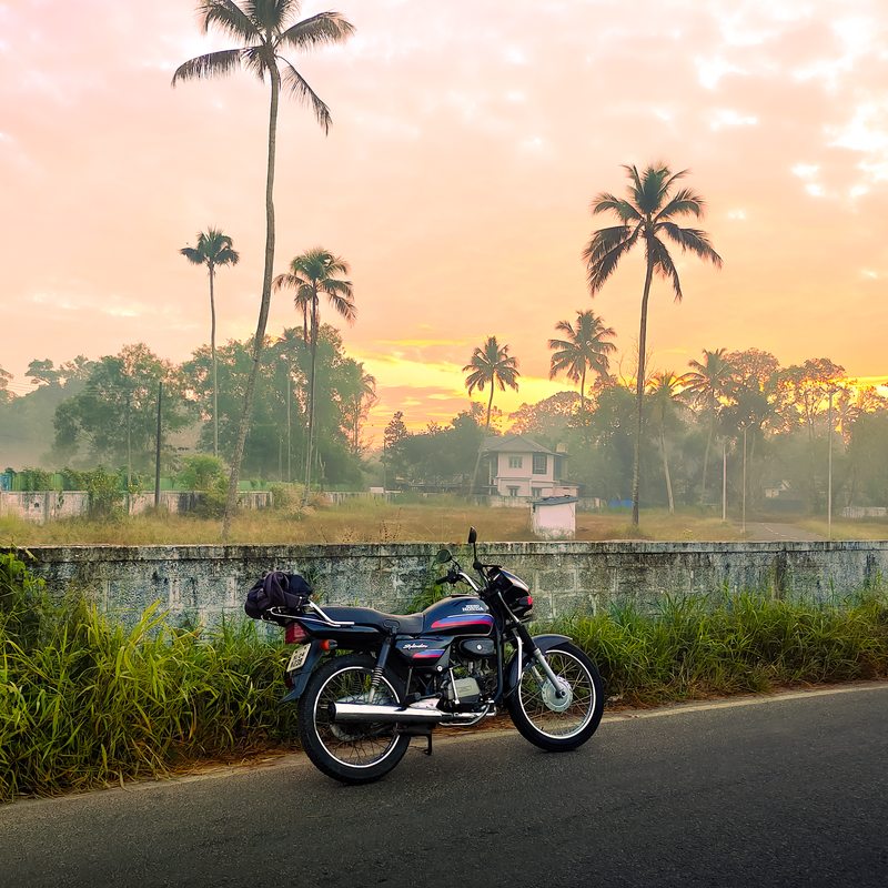 A motorcycle beside a road with coconut palms and a warm sunrise sky